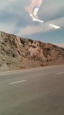 A car is driving on a road. The road is paved with two lanes and a white dividing line. The car is moving from right to left in the frame. To the left of the road is a rocky, barren hillside. The sky is visible above the hill, with clouds. The interior of the car is reflected on the windshield.
