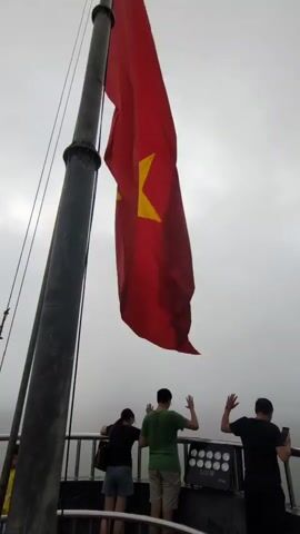 The video captures a low-angle view looking up at a large red flag with a yellow star, likely the flag of Vietnam, on a flagpole. The flag is billowing in the wind against a cloudy sky. Below the flag, on a platform, stand three individuals. One person on the far left, wearing a black shirt and shorts, is leaning on a railing. In the center, a person in a green shirt and shorts stands with their hands raised in a waving gesture. To their right, another person in a black shirt also stands with their hands raised and appears to be holding a phone. A railing separates the individuals from the open space below. In the background, there are lights mounted on the platform.