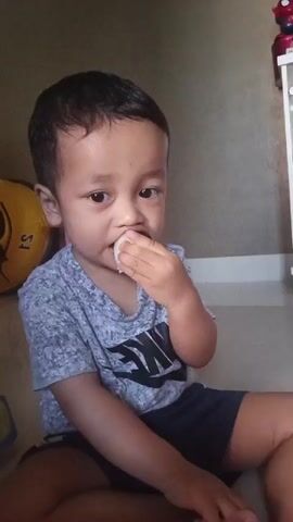 A young boy sits on the floor eating. He is wearing a gray t-shirt and dark shorts. He is holding a piece of food up to his mouth with his right hand. There is a yellow object behind him to the left and a pink drawer unit behind him to the right. The wall behind him is a neutral color.