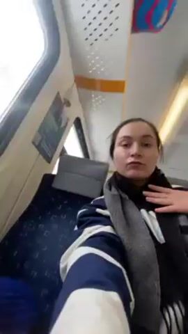 A woman is taking a selfie video on a train. She’s wearing a navy and white striped jacket and a gray scarf. Her hair is pulled back. There’s a small black and brown dog sitting on the seat next to her wearing a blue sweater. The train car has blue seats and windows. The woman looks at the camera and makes a kissy face. Other passengers are visible in the background.