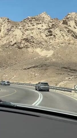 The video begins by showing a silver car driving on a highway through a desert landscape. The car is moving away from the camera, and the road curves to the left. The desert is characterized by rocky, arid mountains under a clear blue sky. The camera is positioned inside another vehicle, looking out through the windshield. White lines mark the lanes of the highway, and a metal guardrail separates the road from the embankment. As the video progresses, the camera continues to follow the silver car. Another white car is seen approaching from the opposite direction. The mountainous terrain remains consistent throughout the brief clip.