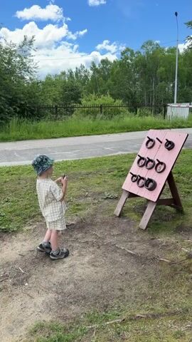 A young child in a light-colored checkered outfit and a patterned hat is playing a ring toss game outdoors. The child stands facing a pink wooden board mounted on an easel, which has several black rings attached to pegs. The child is holding a ring in their right hand, raised as if about to throw it. The child is in the center of the frame, on a dirt and grass area. Behind the game board is a paved area, a grassy embankment, trees, and a blue sky with white clouds. The child moves their arms and body, seemingly excited, and then spreads their arms wide with a smile.