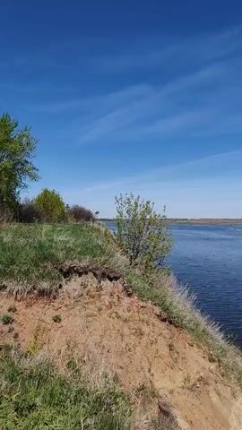 The camera pans from left to right to reveal a river landscape under a blue sky with wispy clouds. In the foreground, a grassy cliff edge overlooks the wide, calm river. A small bush stands on the cliff. In the distance, land can be seen across the river. As the camera pans, a small white boat appears on the river, heading towards the right side of the frame.