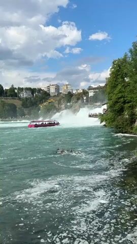 A boat with multiple passengers navigates choppy, turquoise water near a powerful waterfall. Buildings are visible on a hill overlooking the scene, with lush green trees and a partly cloudy sky above. The water is filled with white foam and rapids. The boat appears to be pink and white.