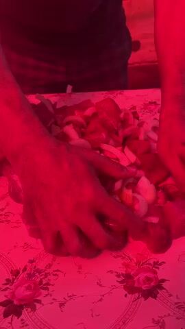 A person's hands are seen preparing a pile of cubed meat and sliced onions on a table covered with a floral tablecloth. The scene is illuminated by a red light, giving everything a reddish hue. The hands are seen mixing the meat and onions.