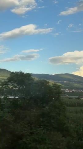 The video shows a landscape with rolling green hills in the background, dotted with houses and what appear to be vineyards. In the foreground, there are fields of crops, with some trees and bushes. The sky is blue with scattered white clouds, some of which are illuminated by the sun. The overall impression is a rural, idyllic setting, possibly in a mountainous region. The camera appears to be moving, suggesting the footage was captured from a vehicle, perhaps a train or car, as it travels through the scenery. There are no people or animals visible in the frame. The lighting suggests it is either morning or late afternoon.