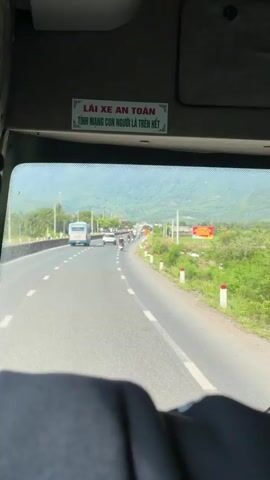 The video shows a view from inside a vehicle, looking out through the windshield. A person's head and shoulders are visible in the foreground, suggesting the viewer is a passenger. The windshield displays a road with traffic, including cars, motorcycles, and a bus. The road is bordered by green fields and hills in the background. There is a sign in Vietnamese on the interior of the vehicle, above the windshield, that reads "LÁI XE AN TOÀN TÍNH MẠNG CON NGƯỜI LÀ TRÊN HẾT" which translates to "DRIVE SAFELY, HUMAN LIFE IS PARAMOUNT."