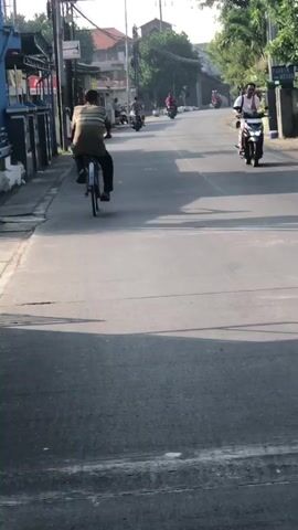 The video shows a street scene with several people riding on motorbikes and bicycles. The road appears to be paved and is lined with buildings and trees. The sun is shining, casting shadows on the road. In the foreground, a person is riding a bicycle. Further down the road, several motorbikes are visible, some carrying multiple passengers. The overall atmosphere is that of a busy street with ongoing activity.