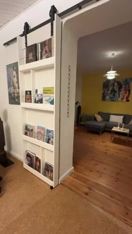 A video pans across an entryway and into a living room. A white, multi-tiered bookshelf with a sliding door is on the left. The bookshelf holds books and magazines. Beyond the entryway, the living room features a yellow accent wall, a gray sectional sofa, a coffee table with candles, a floor lamp, a standing lamp, and a large abstract painting on the wall. The floor is made of hardwood planks. A window with a radiator is visible on the far left wall. The video briefly shows a television and a chest of drawers.