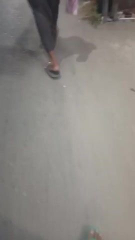 The video shows a person walking on a sidewalk at night. The person is wearing dark pants and sandals. They are carrying a white plastic bag with a pink item inside. The lighting suggests streetlights and possibly headlights from a vehicle. The sidewalk appears to be made of concrete and there is some vegetation and debris on the side. The video is shaky and blurred, indicating movement.