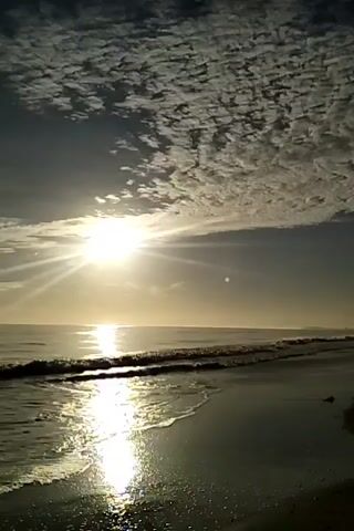The video shows a beach at sunrise or sunset. The sun is shining brightly in the center of the frame, with its rays reflecting off the water. There are scattered clouds in the sky. Gentle waves are lapping at the shore. The sand is dark and wet, reflecting the sunlight.