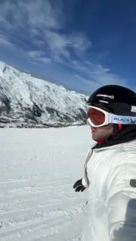Here is a description of the video:
The video shows a person skiing on a snowy mountain. The person is wearing a white ski jacket, black helmet, and goggles. The background shows snow-covered mountains and a blue sky with some clouds. The person points towards the mountains in the background. The camera angle is from the person's perspective, suggesting they are recording themselves while skiing. The snow has lines that appear to be from other skiers.