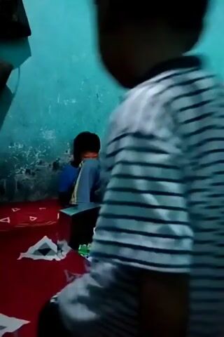 This is a short video clip showing two young boys indoors. The scene appears to be a bedroom, with a red bedspread adorned with a triangular pattern. One boy is seated on the bed, facing away from the camera and towards the right side of the frame. He is wearing a blue shirt and grey pants, with his legs crossed and his arms resting on his lap. In front of him, on the bed, is a cardboard box containing various items, possibly toys or snacks, including some green and white packaging.

The other boy, facing the viewer and positioned in the foreground on the left, is standing. He is wearing a striped t-shirt with horizontal lines in white and dark blue/grey, and dark shorts. His left arm is raised and his fist is clenched, as if he is about to strike or engage in play. His body is angled towards the seated boy. The walls of the room are painted a light blue-green color, and a dark bag hangs from a hook on the wall behind the standing boy.

The lighting in the room is dim, suggesting it might be evening or a poorly lit interior. The overall atmosphere seems playful, with the standing boy's posture indicating an active engagement with the seated boy. There is no visible text in the frame. The context provided indicates the location is Beloh, Indonesia.