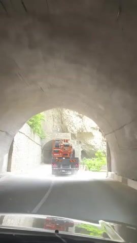 This video captures a journey through a tunnel in Verkhnyaya Balkariya, Russia. The perspective is from inside a vehicle, looking forward.

The primary subject is a truck carrying a large, orange piece of heavy machinery, possibly a loader or excavator, driving into the tunnel. The tunnel is constructed with rough-hewn stone walls and an arched ceiling. Outside the tunnel, the scene is dominated by a steep, rocky mountainside covered in lush green vegetation.

The activity is travel, with the vehicle slowly approaching and then entering the dark tunnel. As the vehicle enters the tunnel, the lighting changes from bright daylight to a dimmer, artificial light. The tunnel walls are wet, suggesting recent rain or moisture. There are lights visible on the tunnel ceiling, illuminating the path ahead. Outside the tunnel, dust or mist is visible in the air near the entrance, potentially from the truck's passage or the environment.

Notable details include the rough texture of the tunnel's stone construction and the natural, rugged beauty of the surrounding mountain landscape. The time of day appears to be daytime, given the bright light outside the tunnel. The weather seems to be damp. The presence of the truck and heavy machinery suggests potential construction or maintenance work in the area. The overall mood is one of transit through a dramatic and somewhat enclosed natural environment. There is no visible text in the media.