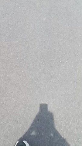 The video shows a first-person perspective of someone walking on a paved path. The path is gray asphalt with a white line on the right side. There is grass and trees on both sides of the path. The sky is bright and appears to be daytime. The person's shadow is visible on the path in the beginning of the video. There are streetlights visible in the distance.