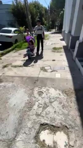 A woman pushes a child in a purple tricycle down a sidewalk. The woman is wearing a white t-shirt, dark pants, and a white bag hangs from her shoulder. The tricycle has a purple sunshade. A white car is parked on the street. The sidewalk is concrete with some patches of weeds growing through cracks. Buildings and trees line the street. The day is sunny.