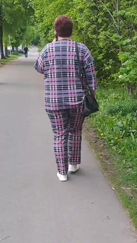 A woman with short red hair is walking away from the camera on an asphalt path in a park. She is wearing a matching plaid set in shades of gray, pink, and white, and white sneakers. She carries a black purse over her shoulder and is talking on her cell phone. The path is lined with trees, and grass and dandelions grow on the side. Other people and cars are visible in the distance.
