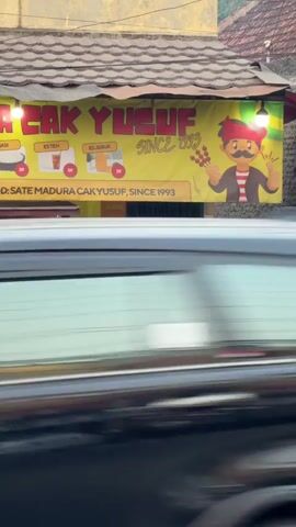 The video captures an outdoor food stall operated by a man identified as Cak Yusuf. He is wearing a traditional red fez and a striped shirt, actively fanning a grill with a woven fan, generating smoke. The stall's signage, in yellow, prominently displays "CAK YUSUF SINCE 1993" and includes illustrations of drinks and satay. To the left, another person stands behind a counter, and there are bags and some produce, possibly cabbage, near the grill. The stall appears to be situated by a road, with blurred vehicles passing in the foreground, indicating a street-side location. The overall atmosphere suggests a bustling, casual food service environment.