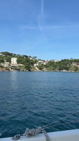 The video opens with a serene view of a coastline dotted with white houses and lush greenery, as seen from a boat on a calm, blue sea. The sky is a clear blue with faint contrails. The camera slowly pans to the left, revealing more of the coastline and the houses that are nestled into the hillside. Eventually, a white sailboat comes into view in front of the land mass. The perspective remains steady as the boat appears to be moving slowly through the water. The overall scene is peaceful and picturesque.