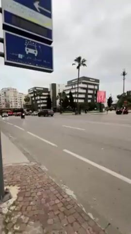 The video shows a street scene in a city. Cars and motorcycles are driving on a wide, multi-lane road with palm trees lining the median. Buildings are visible in the background, including a modern, glass-fronted building. There are also street signs and light poles. The sky is overcast.