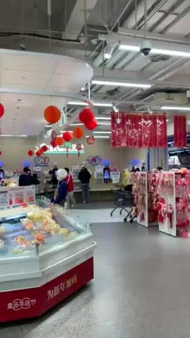 The video shows the interior of a brightly lit supermarket adorned with red and orange decorations, indicating a festive occasion such as Chinese New Year. Various people are visible, including women in casual attire, a man pushing a shopping cart with a child inside, and shoppers browsing the aisles. The supermarket features displays of food items in refrigerated cases and decorative items on shelves. The overall atmosphere suggests a bustling shopping environment, potentially during a holiday season.