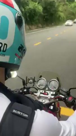 A person wearing a blue and white helmet and a black Burton backpack is riding a red motorcycle on a two-lane road. There are other cars and motorcycles on the road. On the right-hand side of the road there are green hills and trees. There is also a sign indicating “Son Tra”. The weather appears to be overcast.