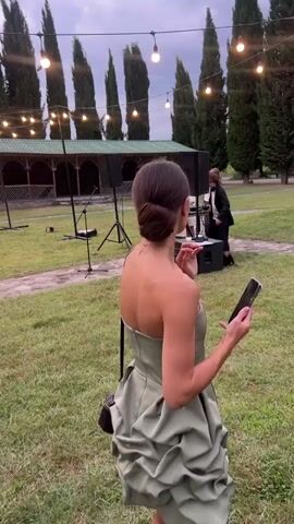 The video shows an outdoor event with a woman in a green dress in the foreground, holding a phone. In the background, there's a man in a white shirt and black pants, and another man in a white shirt and beige pants. There are tables and chairs set up for dining, string lights overhead, and tall trees surrounding the area. The event appears to be in the evening.