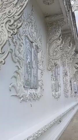The video pans across the exterior of a white building with intricate white relief carvings and decorations. The building appears to be a temple or a similar ornate structure. The carvings are detailed and elaborate, featuring swirling patterns and stylized figures. The camera moves from left to right, revealing multiple windows with decorative frames. The building's wall is pristine white, and the carvings are also white, creating a monochromatic aesthetic. The ground in front of the building is paved with light-colored tiles. The sky is visible in the background, with some clouds against a blue backdrop. The camera continues to pan, showing more of the building's facade and the surrounding architecture, which also features similar white ornate designs. The overall impression is one of elaborate craftsmanship and artistic detail in a bright, white environment.