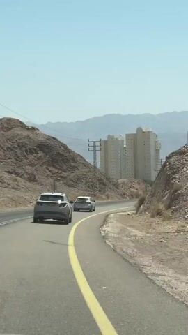 The video shows a view from a car driving on a paved road through a dry, mountainous landscape. Two cars are ahead on the road, one gray SUV and a lighter colored car behind it. In the background, there are several tall, light-colored buildings, likely hotels or apartment complexes, situated near the sea. Mountains are visible in the distance. A tall cell tower stands on the right side of the road. The sky is clear and blue. The sun is shining brightly, casting shadows. The road has a yellow line dividing the lanes. The general environment appears arid and desert-like.