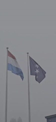 The video shows two flags waving in the wind against a gray sky. One flag is the flag of Luxembourg, with horizontal stripes of red, white, and light blue. The other flag is blue with three white stars and the word "ROTAREX" below the stars. The flags are mounted on tall, silver poles.