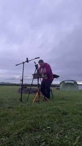 A man wearing a purple hoodie is playing a steel drum in a grassy field. There are microphones set up around him. In the background, there are tents and a campfire where a group of people are gathered. The sky is overcast.