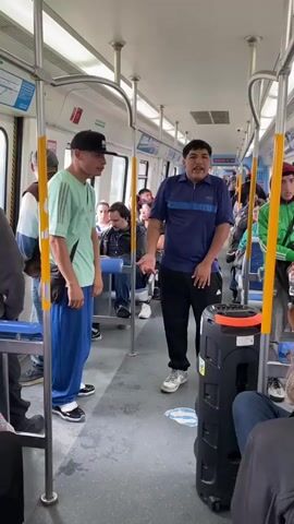 The video shows the interior of a clean, well-lit train or subway car, likely in Piñeyro, Argentina. Two men are standing in the foreground in the aisle. On the right, a man wearing a blue polo shirt and black pants is speaking or singing energetically, gesturing with his hands. He stands next to a large, black portable speaker on wheels, which has an orange top. On the left, a man in a light green t-shirt, loose blue pants, and a black baseball cap is listening intently, facing the speaker. He wears a small black bag across his chest. Numerous other passengers are visible in the background, some seated on blue seats and others standing, observing the activity. The train car has yellow handrails, white walls, and a grey floor with a visible white sticker or marking near the speaker. The scene depicts an impromptu performance or address occurring during the daytime within the public transport vehicle.