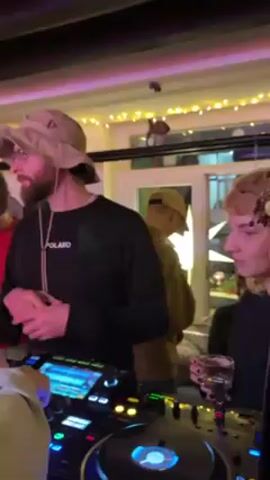 Here’s a concise description of the video:

The video takes place indoors, seemingly at a party. A bearded man wearing a beige bucket hat, glasses, and a black long-sleeved shirt with "POLAKO" printed on it stands in front of DJ equipment. He gestures toward his chest with both hands. He is surrounded by other people, some of whom are dancing or looking on. Fairy lights are strung across the background. The DJ equipment is illuminated with bright lights, and music plays.
