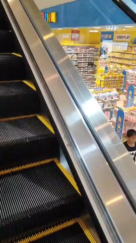 The video shows an escalator in what appears to be a toy store. The escalator steps are black with yellow safety lines. The handrail is silver and metallic. In the background, shelves are stocked with toys, including LEGO products. A sign with colorful letters that seems to spell "KIDZANION" is visible on the upper wall. The perspective is from someone standing on the escalator, looking upwards and to the right. The lighting is bright, typical of a retail environment. There are no people clearly visible on the escalator or in the immediate vicinity. The video is short, lasting only a few seconds, and captures the upward movement of the escalator.