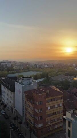 The video shows an elevated view of a city at sunset or sunrise. The sky is a mix of orange and light blue, with the sun low on the horizon. The cityscape includes a variety of buildings with different roof styles, including flat, tiled, and gabled. Trees are scattered throughout the city. A building with a red cross symbol is visible. Cars are parked along a street. The camera pans slowly across the scene.