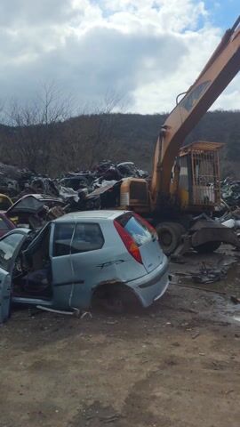 The video shows a junkyard. There are many wrecked cars, trucks, and tires piled up. There is an orange excavator and a tractor. The sky is blue with white clouds. The junkyard is on a dirt lot, and there is a small hill in the background.