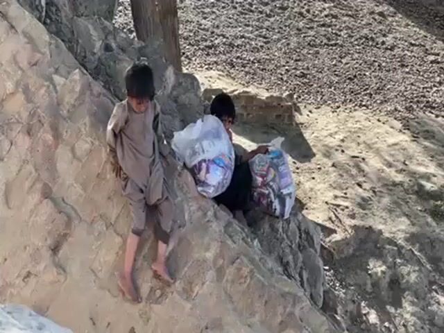 Here's a detailed description of the video:

**Scene:** The video appears to be shot outdoors, likely in a rural or less developed area. The ground is a mix of dirt, dry vegetation, and what looks like a constructed stone terrace or wall.

**People:** Two young boys are visible in the frame. One is sitting on the wall/terrace, while the other is standing on the lower portion of it. They both seem to be barefoot.

**Clothing:**
*   One boy is wearing a grayish-tan, loose-fitting traditional garment.
*   The second boy is wearing a dark-colored outfit.

**Objects:**
*   Two large, clear plastic bags are visible. They appear to be full of various packaged food items, possibly candy or snacks.

**Actions and Interactions:**
*   The boys are positioned on the rocky structure, appearing to be handling or carrying the bags of goods.
*   The boy who is sitting is looking at the bags.

**Environment:**
*   The background is somewhat blurred, but the terrain looks like a rocky or dirt hill with scattered vegetation.
*   There is a tree visible in the background near the stone structure.

In summary, the video shows two young boys, possibly in a rural area, sitting and standing on a rock wall, each carrying large bags filled with what seems to be food items. The scene suggests they may be involved in distribution or collection of these items.