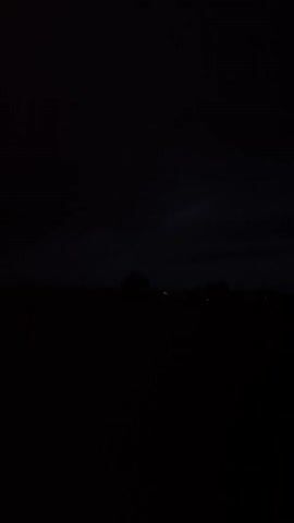 This is a dark video, likely taken at night or in very low light conditions. The foreground is almost entirely black, with no discernible objects or details. In the middle ground, there is a dark silhouette of what appears to be trees or vegetation, with some faint, distant lights visible. The sky is also dark and overcast, with no stars or moon visible. The video is still, with no movement or action captured. It is difficult to determine the exact location or time of day due to the lack of light and detail.