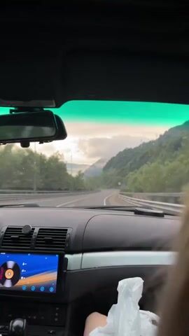 The video shows the interior of a car driving on a road with a mountainous landscape. The dashboard is visible, with a screen displaying a music interface and what appears to be a vinyl record graphic. The windshield offers a view of a winding road ahead, flanked by lush green hills and trees. The sky is overcast with clouds, and the overall mood suggests a scenic drive. There is a person's leg visible in the lower right corner of the frame, and a plastic bag on the passenger seat.