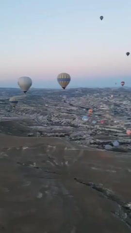 This is a first-person perspective video from a hot air balloon. Numerous other hot air balloons are visible in the sky, scattered across a vast, mountainous landscape. The sun appears to be rising or setting, casting a soft light on the scene. The landscape below is characterized by rocky, eroded terrain, with some signs of human settlement or structures visible in the distance. The person recording the video occasionally speaks, possibly to someone else in the balloon or to the camera.