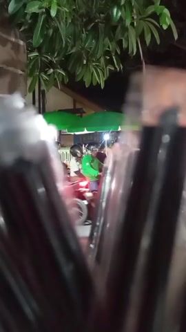 The video captures a nighttime scene with the foreground blurred by dark, cylindrical objects, possibly packaging or containers. In the background, a group of people are visible under green umbrellas, suggesting an outdoor market or food stalls. There are lights illuminating the area, and some trees with green leaves are present. One person appears to be on a motorcycle. The interaction between the people is not clear due to the distance and blurriness. The overall atmosphere is that of a lively, open-air gathering in the evening.