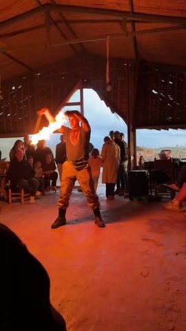A young man is doing a fire dance in a room with a pitched roof. The room has a rough, rustic feel. He is wearing a dark t-shirt, tan pants, and boots. He is holding a flaming torch in each hand and spinning them around his body in a rhythmic motion. A small crowd of people are watching him, some seated on chairs and others standing near the windows. The windows look out onto a cloudy landscape. A speaker is visible on the floor beside the standing people. The room has a red tint to the floor, likely from the firelight. The light is dim and the room is filled with smoke.