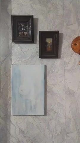 The video pans across a wall adorned with three framed pictures and an unframed canvas. The wall has a textured, light-colored wallpaper with a subtle pattern.

The top left picture is a small, dark-framed landscape depicting a lone tree with bare branches silhouetted against a bright sky, possibly a sunrise or sunset, with a body of water in the foreground.

To the right of that, also in a dark frame, is another small landscape. This one appears to show a body of water reflecting a colorful sunset or sunrise with a few silhouetted trees or structures on the horizon.

Below these two framed pictures is a larger, unframed canvas. The canvas is mostly white with washes of light blue and possibly some light gray or white abstract markings. In the upper central area of this canvas, there is a faint, circular shape.

On the far right edge of the frame, a small portion of a round, wooden object is visible, possibly a clock or a decorative element.