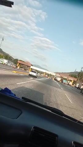 The video shows a car approaching a toll booth. The booth has several lanes, and cars and trucks are moving through them. The sky is blue with some clouds, and mountains can be seen in the background. The camera is positioned inside the car.