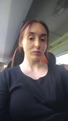 The video shows a woman on a bus. She is wearing a black long-sleeved shirt. Her reddish hair is pulled back. The seats are gray and orange. The bus is moving, as the background seen through the window is blurred.