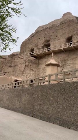 The camera pans slowly upwards to reveal a cliff face that has been carved to include several structures, all in a sandy brown color. Along the middle section of the cliff face are two tiers of wooden walkways with railings, each leading to openings that look like caves. Below this, closer to the ground, is a small building, also painted the same sandy brown. There is a fence in front of the building, and a set of steps is visible leading up to the building, with a fence lining the steps. The area is partially paved. The sky is overcast. There is some green foliage visible to the left of the frame.