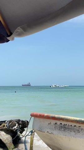 The view from under a sunshade shows a beach scene with a calm, aquamarine sea under a clear blue sky. A white and red fishing boat, labeled "El Arca de Noe" in black lettering, sits in the foreground, partially obscured by the sunshade. In the background, several people are swimming in the water. Further out to sea, two boats are visible, one a small speedboat, and the other a large cargo ship. The overall scene is peaceful and scenic.