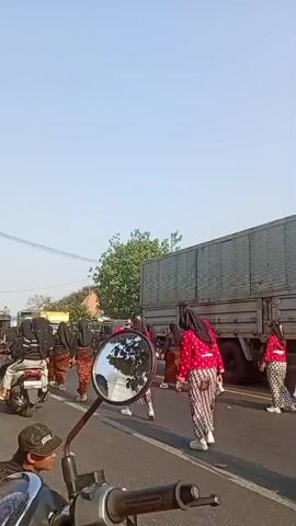 A group of people dressed in traditional clothing are walking on a road next to a large truck. Some are wearing red tops and patterned skirts, while others are wearing white tops and patterned sashes. The individuals appear to be participating in some sort of procession or cultural event. The scene is lit by natural daylight, and the sky is clear and blue. A motorcycle is parked in the foreground, partially obscuring the view.