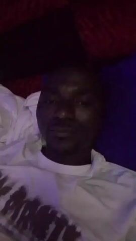 The video shows a person lying down and looking into the camera. They are wearing a white t-shirt with a design on it. The person is in a dimly lit room with what appears to be a red patterned surface behind them.  There is also a white pillow and a blue surface visible. The camera remains stationary throughout the video.
