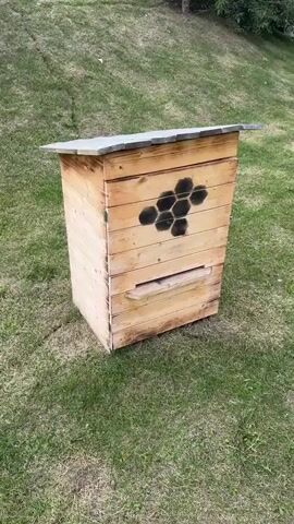 The video shows a wooden beehive with a hexagonal honeycomb design painted in black on its front. The beehive is positioned outdoors on a grassy area, with the grass appearing somewhat dry and patchy. The beehive is made of horizontal wooden planks and has a pitched roof covered with what appears to be a gray material. There is an entrance opening on the lower front of the beehive, with a small wooden landing board extending outwards. The video is a close-up view of the beehive, with the camera slowly zooming in and panning slightly. There are no people or animals visible in the video. The lighting suggests it is daytime, possibly overcast.